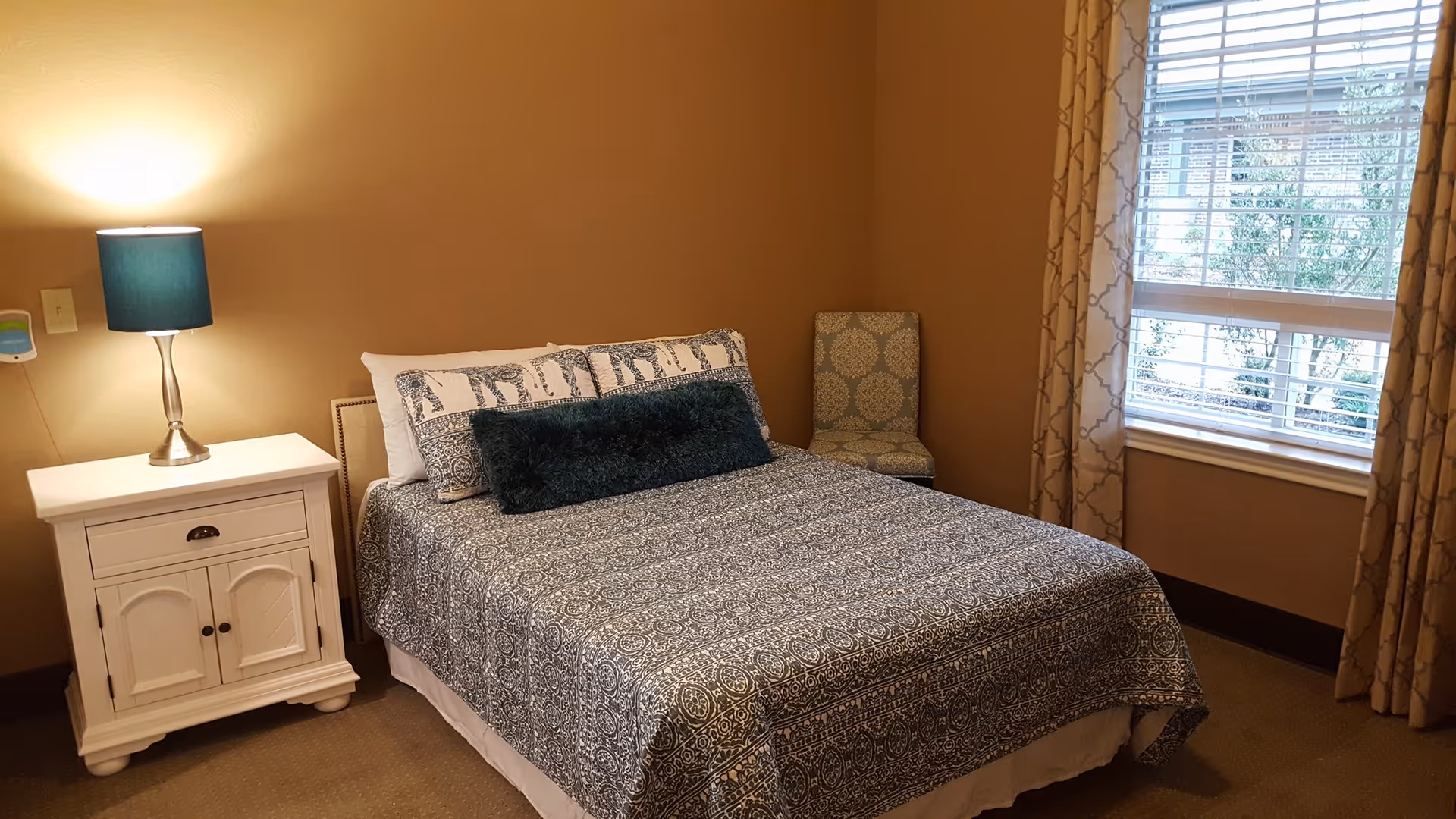 A cozy bedroom with a neatly made bed featuring patterned bedding and multiple pillows. To the left of the bed is a white nightstand with a blue lamp on it. A patterned chair is placed in the corner near a window with beige curtains and white blinds, allowing natural light to enter the room.