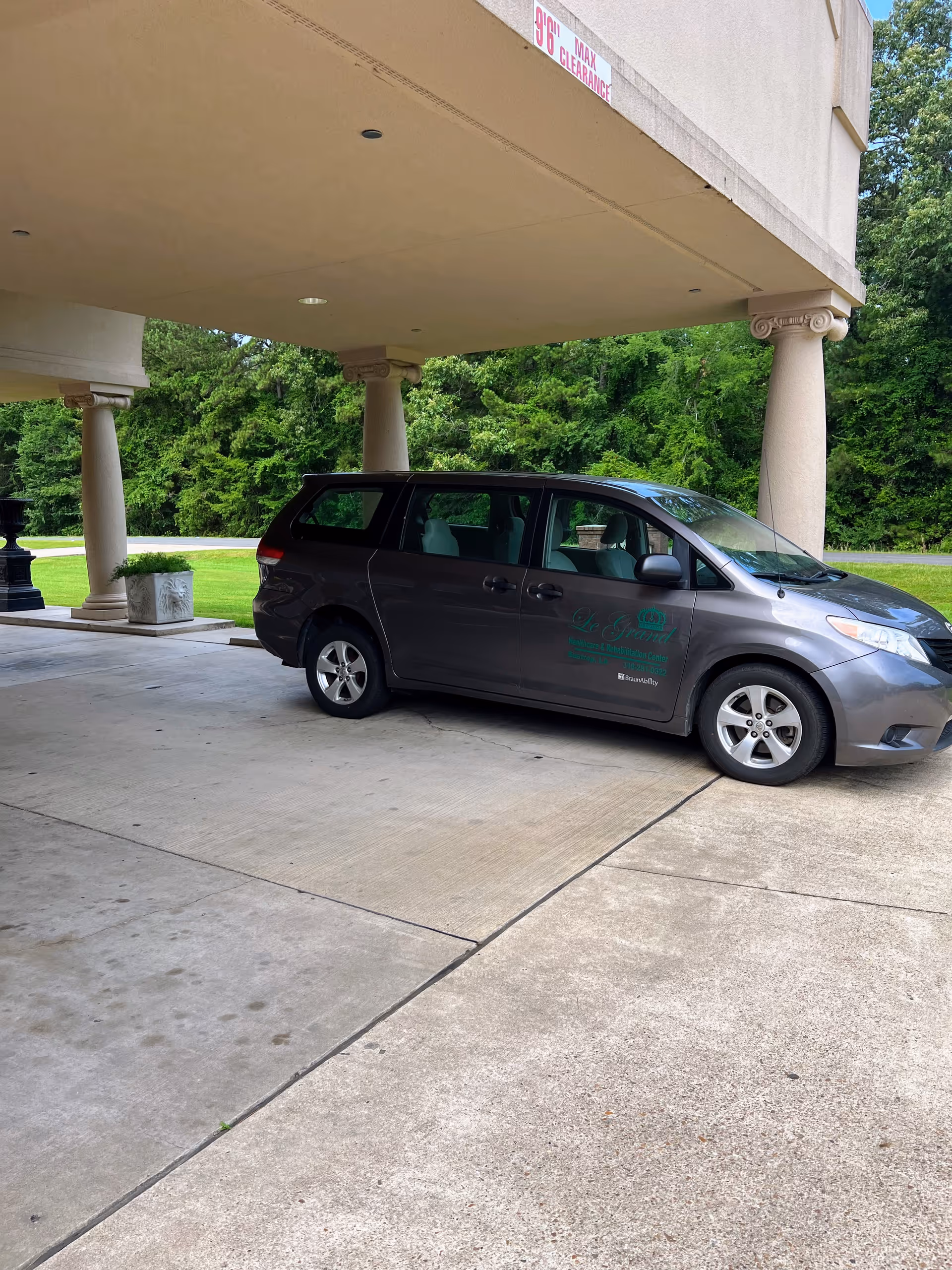A gray minivan parked under a covered driveway with beige columns. The minivan has the logo and name 'LeGrand Health Care & Rehabilitation Center' on its side. In the background, there is green grass and trees.