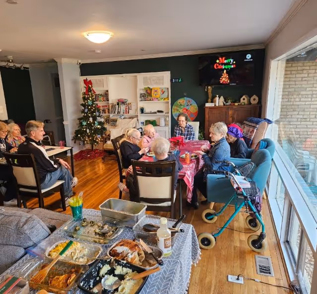 A group of elderly people sitting around a table covered with a red tablecloth, enjoying a meal together in a cozy room decorated with a Christmas tree and holiday decorations. There is a large window letting in natural light, a television on the wall displaying a Merry Christmas message, and a buffet table with various dishes in the foreground.