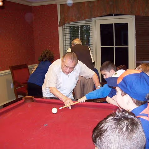 An elderly man playing pool with a group of children gathered around a red pool table in a room with red wallpaper and a window with blinds.