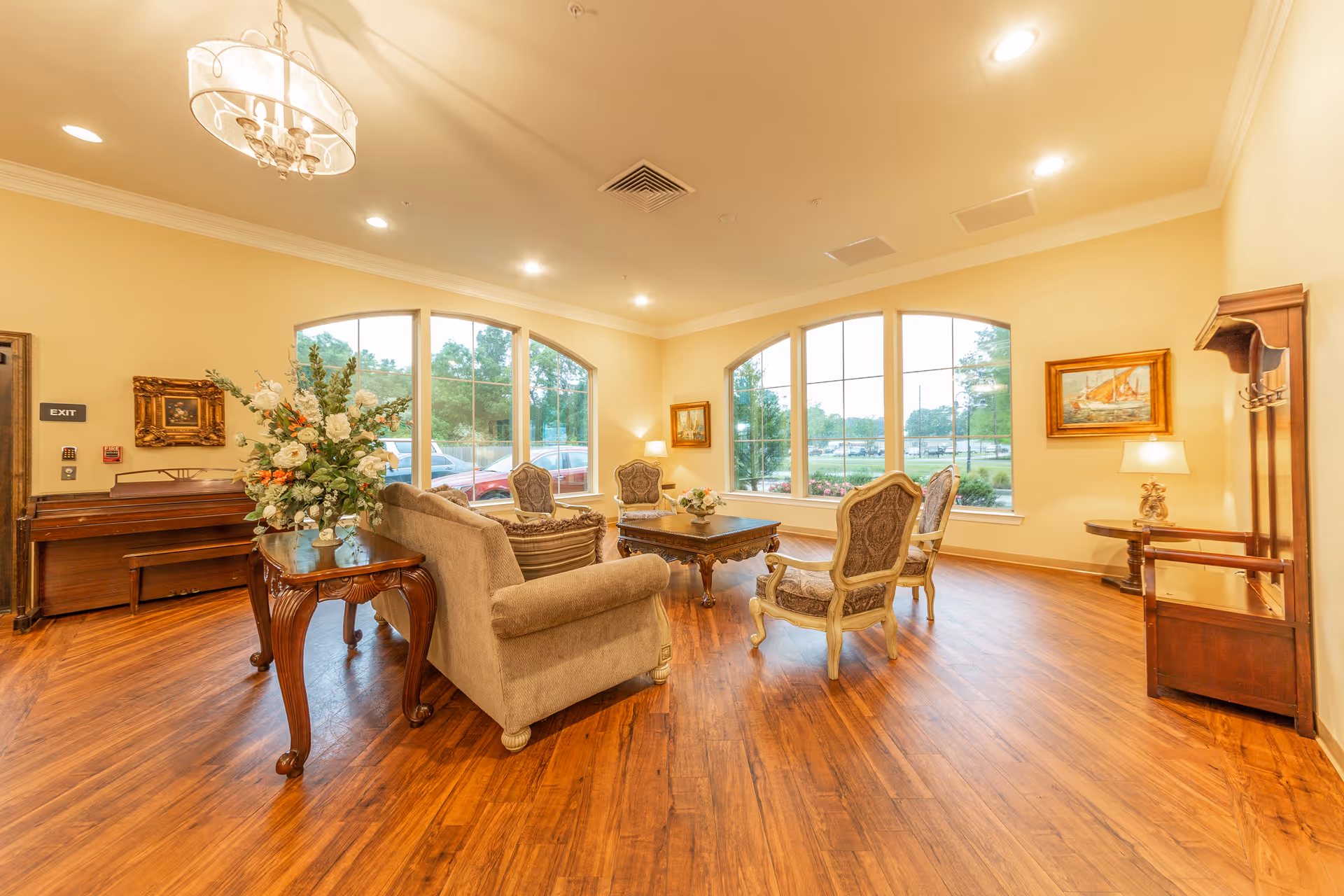 Well-lit sitting area with sofas, upholstered armchairs, a coffee table, piano, and large arched windows overlooking greenery.