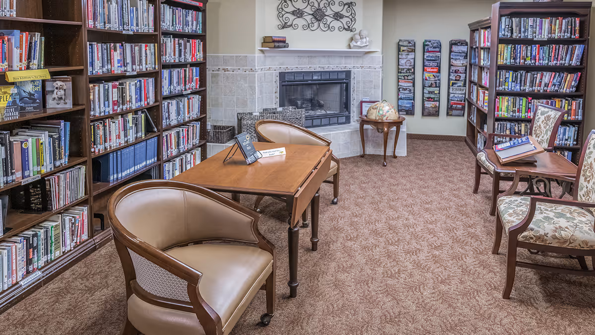 A cozy library room with bookshelves filled with books on both sides, a wooden table with two chairs in the center, a fireplace with a tiled surround at the back, a small round table with a globe on it, and magazine racks on the wall.