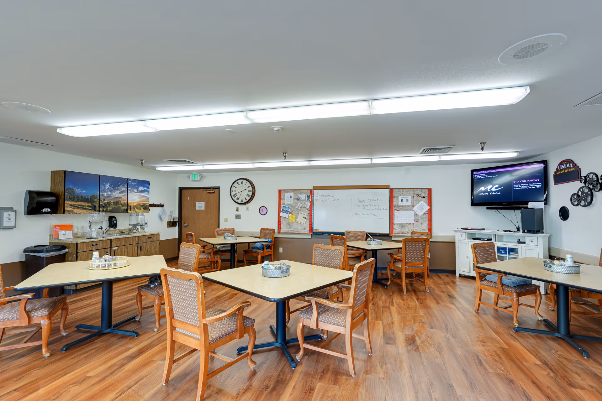 Bright common dining/activity room with multiple tables and chairs, a TV, wall clock, whiteboard and a kitchenette area.