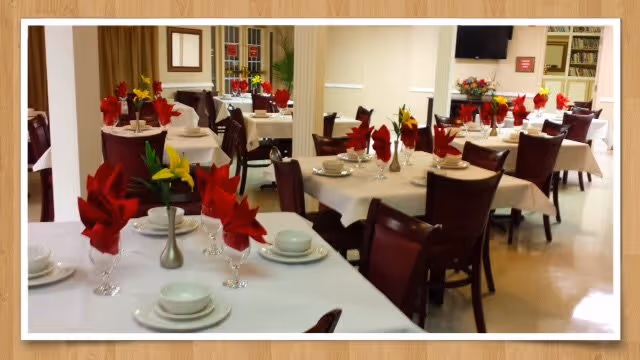 A dining room set up with multiple tables covered with white tablecloths, each table adorned with red and yellow folded napkins in glasses, white plates, bowls, and silverware. The room has wooden chairs with dark cushions and a bright, clean atmosphere with windows and a television mounted on the wall.