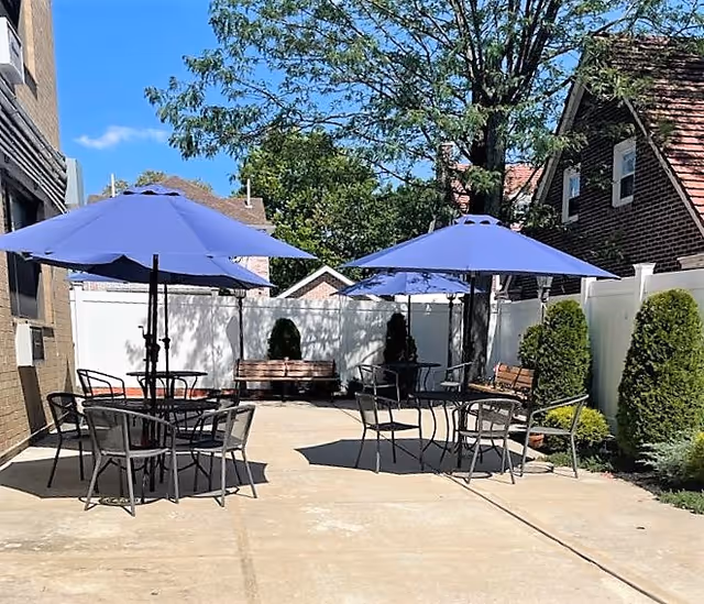 Outdoor patio area with several black metal tables and chairs, each table shaded by a blue umbrella. There are wooden benches along the white fence, with green shrubs and trees surrounding the space under a clear blue sky.
