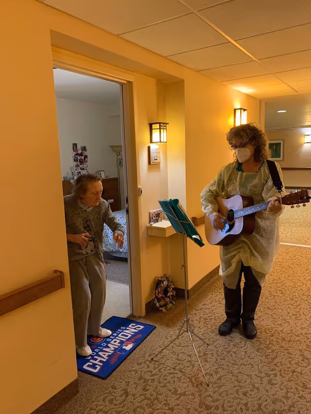 An elderly woman stands in the doorway of her room inside a senior living facility, watching a woman wearing a mask and protective gown play an acoustic guitar in the hallway. The hallway is warmly lit with wall sconces, and there is a music stand holding sheet music in front of the guitarist. A blue doormat with 'World Series Champions' text is visible at the doorway.