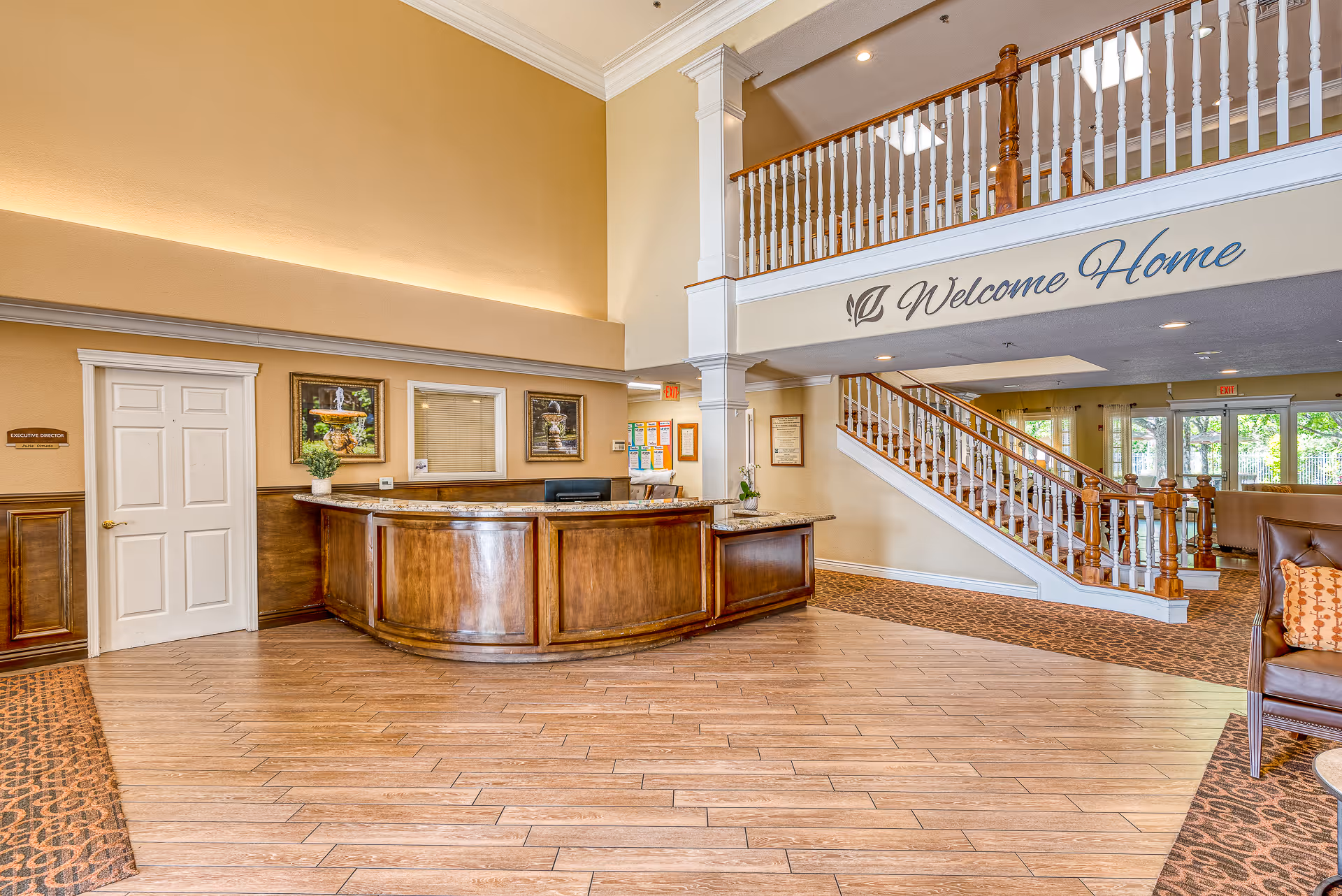 Lobby of a senior living facility with a curved wooden reception desk, a staircase, and a 'Welcome Home' sign on the upper wall.