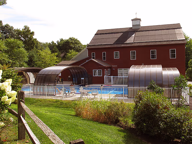 Outdoor view of a red building with solar panels on the roof, surrounded by greenery and trees. In front of the building is a fenced swimming pool with two transparent curved pool enclosures. There is a wooden fence and flowering bushes in the foreground.
