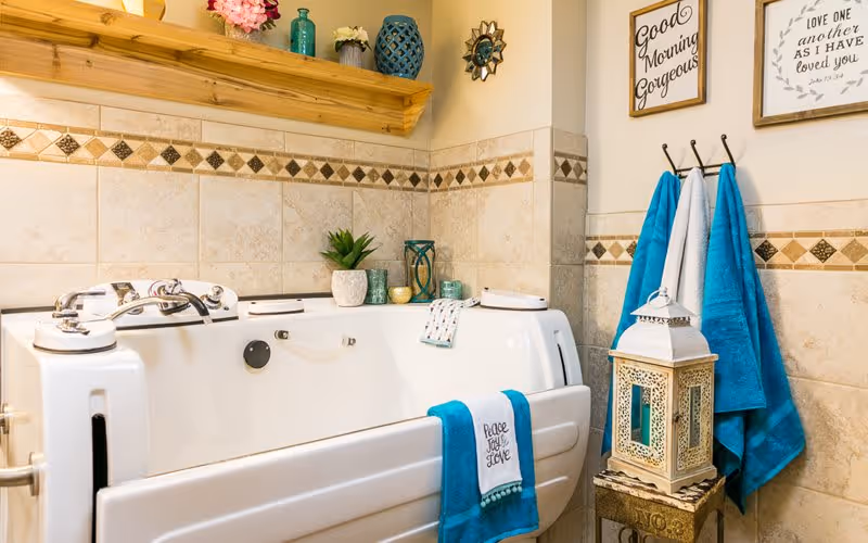 A bathroom with a white walk-in bathtub featuring silver faucets and a blue towel hanging on the side with the words 'Peace Joy Love'. The walls are tiled with beige tiles and a decorative diamond pattern border. Above the tub is a wooden shelf with decorative items including vases and flowers. On the right wall, there are two framed signs with positive messages and three towels hanging on hooks. A decorative lantern sits on a small table next to the tub.