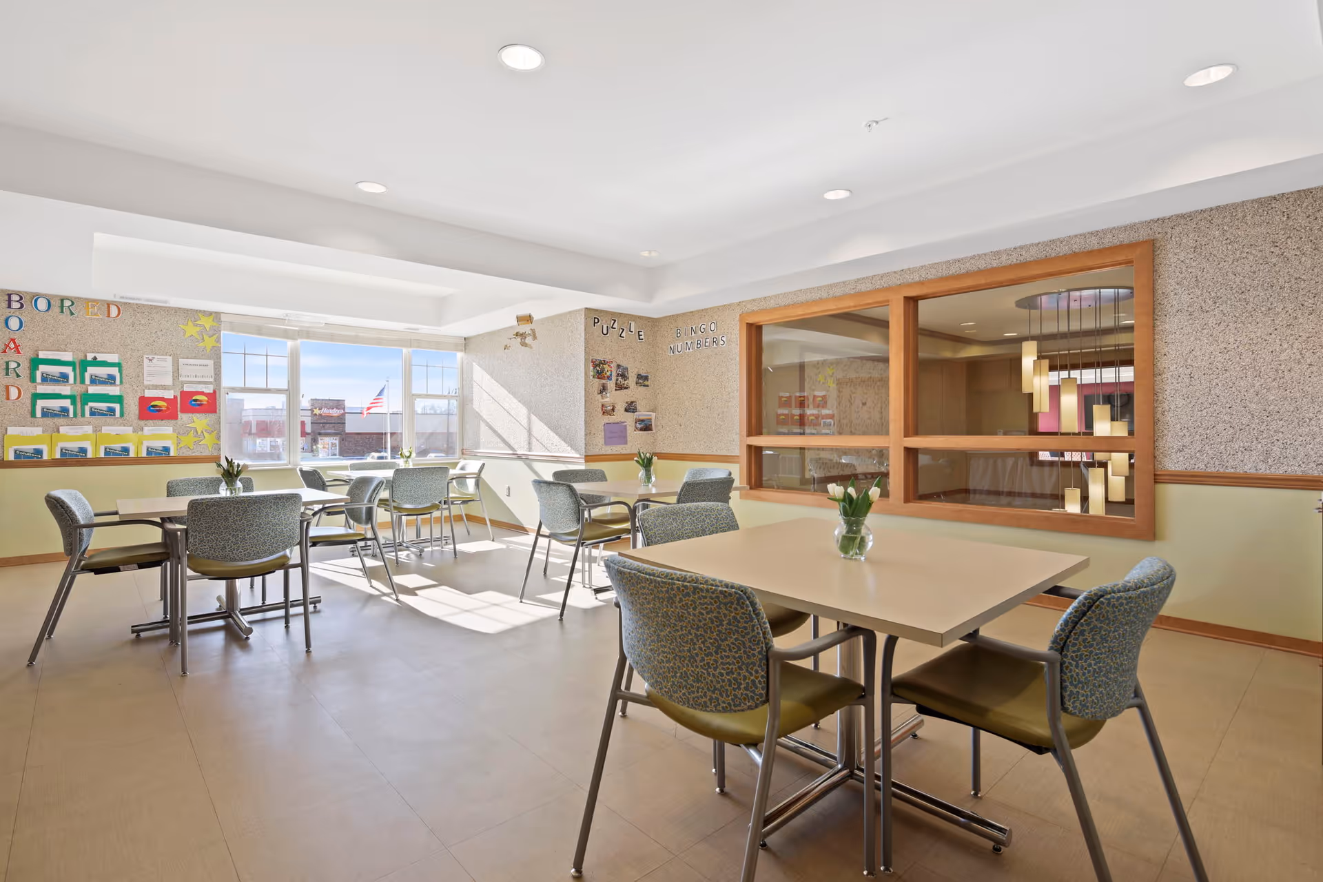 A bright and clean common room with several tables and chairs arranged for group activities or dining. The room has large windows letting in natural light, bulletin boards on the walls with various notices and decorations, and a glass window looking into another room with hanging pendant lights.