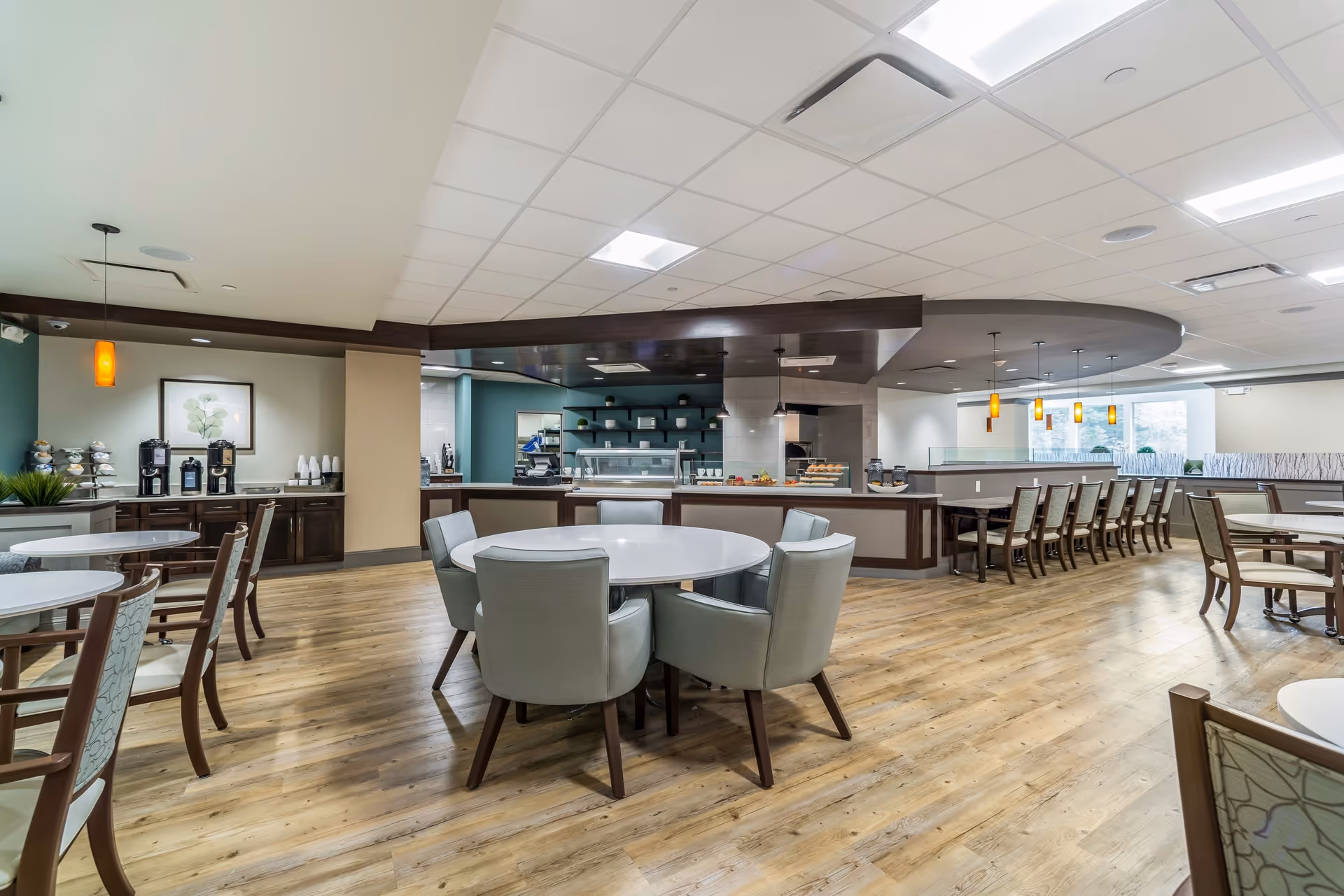 A spacious and modern dining area in Gambrill Gardens Senior Apartments featuring round tables with cushioned chairs, wooden flooring, pendant lighting, and a serving counter with food displays and coffee machines in the background.