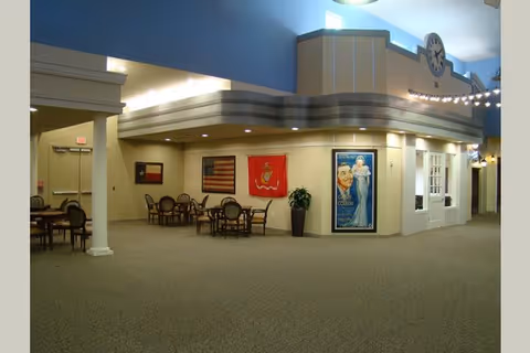 Interior view of a senior living facility common area with several round tables and chairs arranged along the walls. The walls display framed flags and a vintage-style poster. The ceiling is high with a blue upper section and a clock mounted on a decorative structure. The carpeted floor and soft lighting create a welcoming atmosphere.