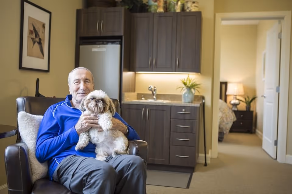 An elderly man wearing a blue jacket sits in a brown leather armchair holding a small fluffy dog. Behind him is a kitchenette with dark wood cabinets, a small sink, and a refrigerator. To the right, an open doorway leads to a bedroom with a nightstand, lamp, and bed partially visible.