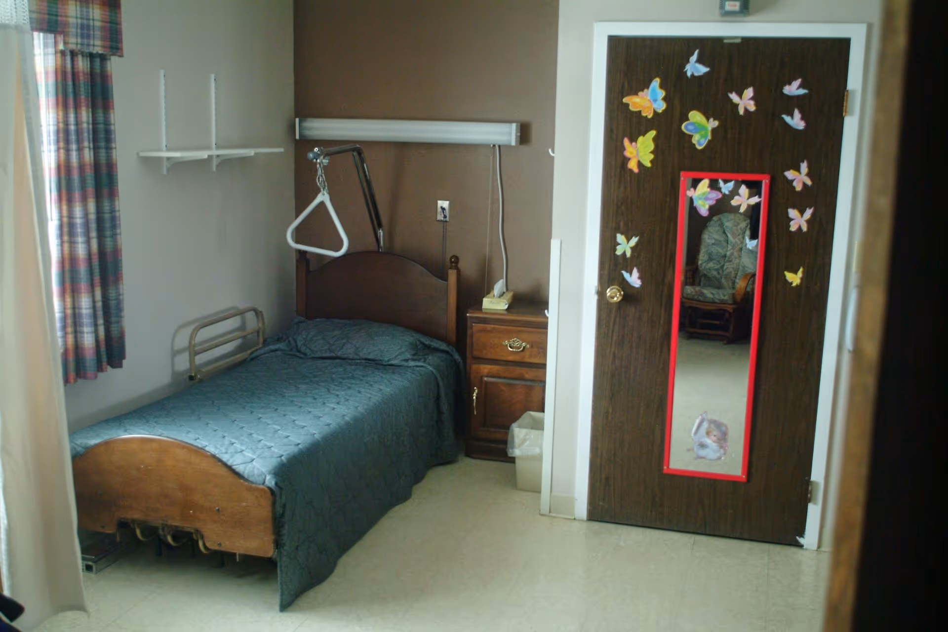 A small bedroom in a senior care facility with a single bed covered in a dark green quilt. There is a wooden headboard and a trapeze bar above the bed. Next to the bed is a wooden nightstand with a telephone and a trash bin underneath. The door has a full-length mirror bordered in red and decorated with colorful butterfly stickers. A cushioned chair is visible through the reflection in the mirror. The room has plaid curtains on the window.