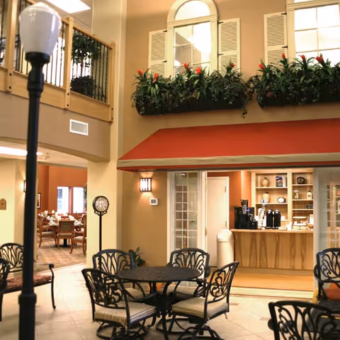 Indoor seating area with black metal tables and chairs with cushions, a small coffee or refreshment counter with coffee machines, and a dining room visible in the background. The area has a high ceiling with windows, decorative plants, and a red awning above the counter.