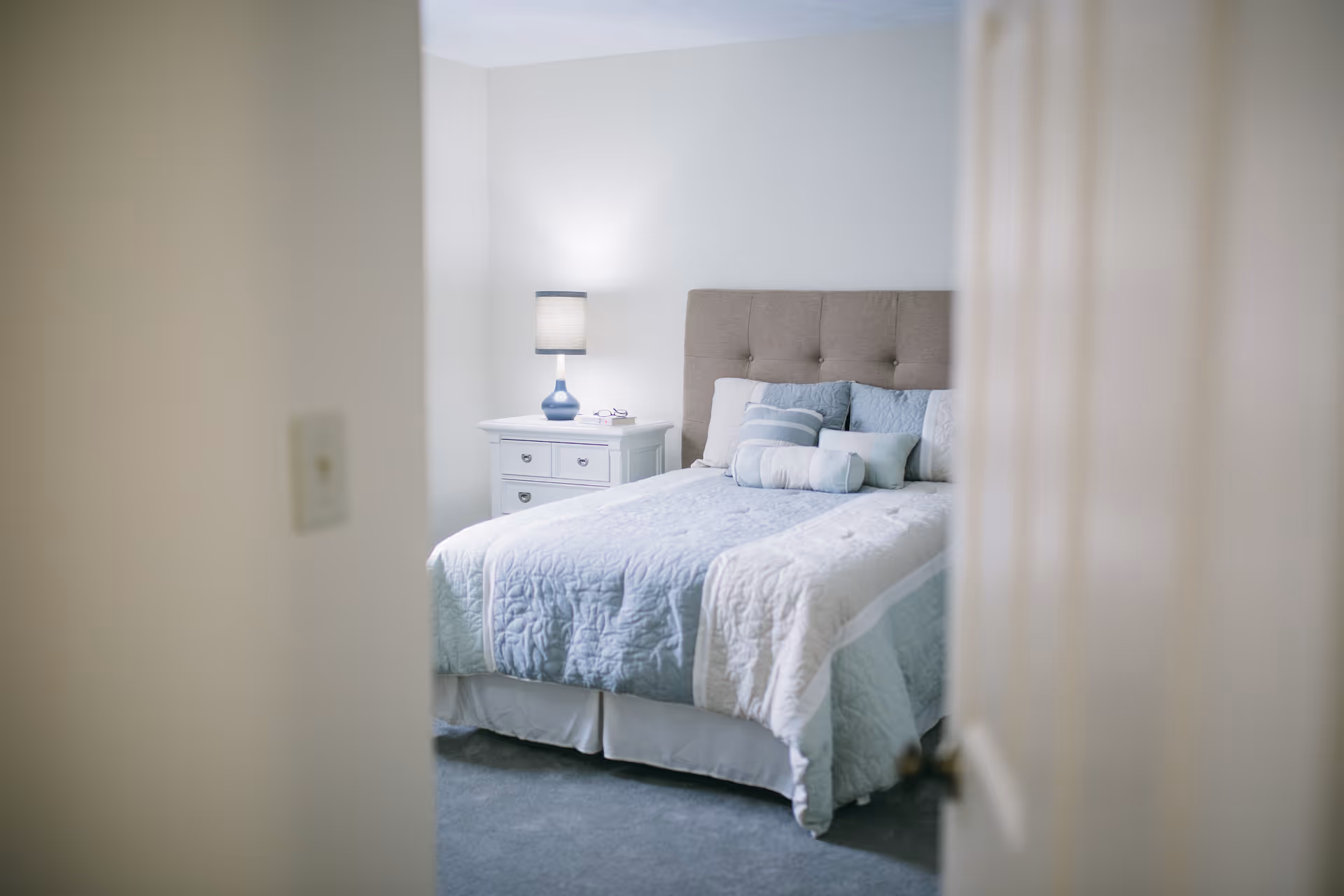 View through a partially open door into a bedroom with a neatly made bed featuring a tufted headboard, light blue and white bedding, and several pillows. A white nightstand with drawers holds a blue lamp and a pair of glasses. The room has light-colored walls and carpeted floor.
