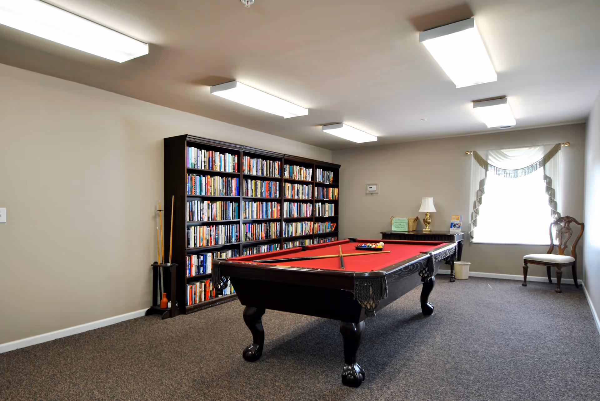A bright interior recreation room featuring a red-felt pool table, a large bookshelf, and seating by a window.