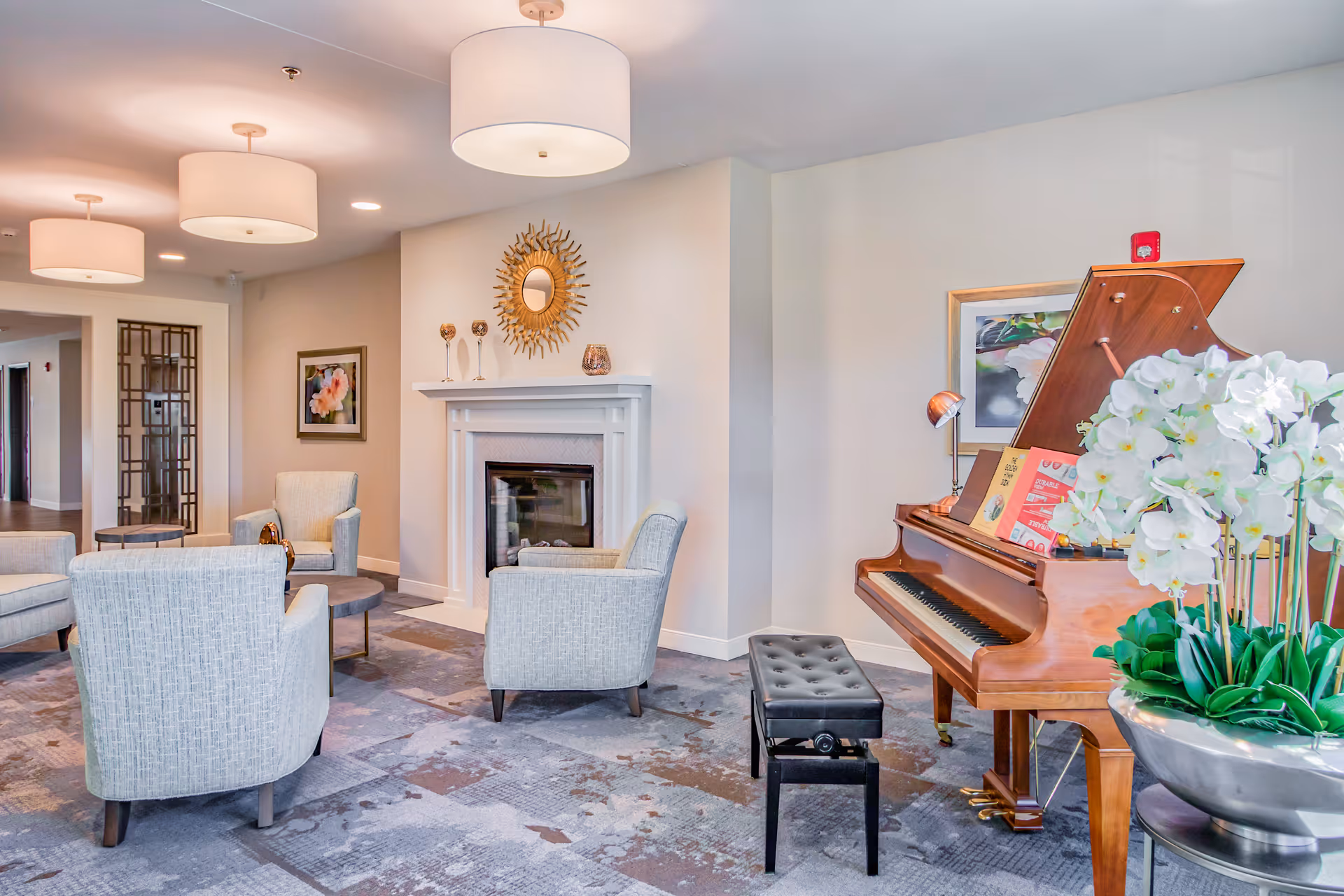 A bright and spacious senior living facility common area featuring a wooden grand piano with a black cushioned bench, a large silver vase with white orchids, a white fireplace with decorative items on the mantel, four light gray upholstered armchairs arranged around small round tables, modern ceiling lights, and framed artwork on the walls.