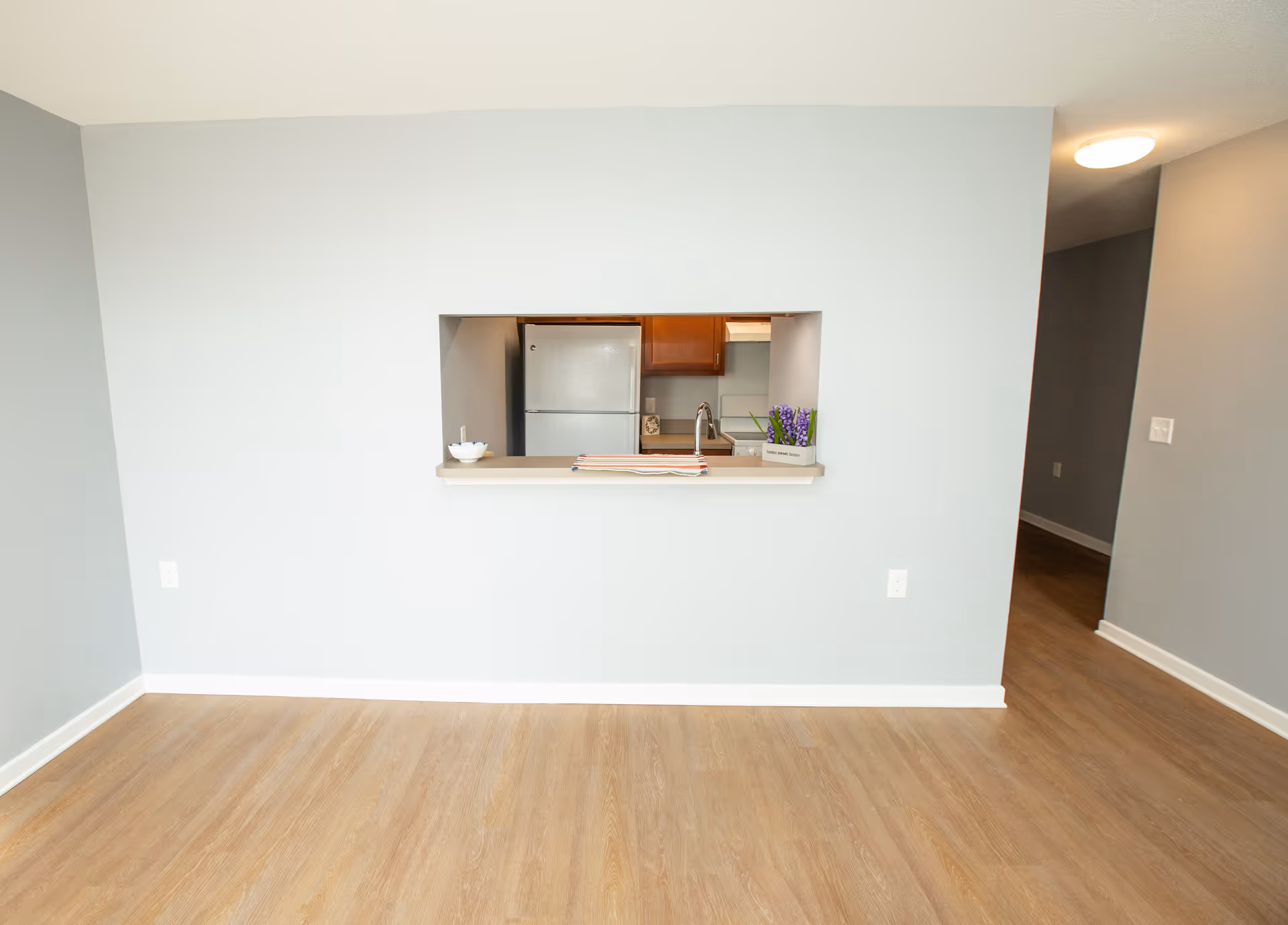 View of an empty room with light wood flooring and light gray walls. There is a rectangular pass-through window in the wall showing a kitchen area with a refrigerator, wooden cabinets, and a sink. A small bowl, folded cloth, and a small potted plant with purple flowers are on the pass-through counter.