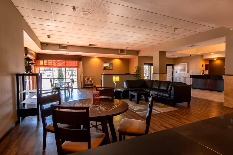Spacious senior living common area with a round table and chairs, a leather sectional sofa, and a reception desk in the background.