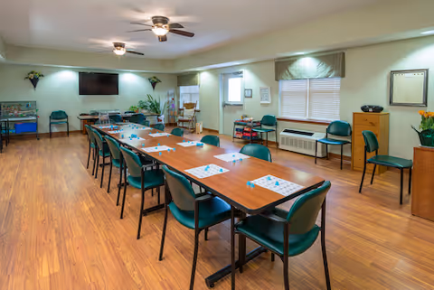 A spacious room with a long rectangular table surrounded by green chairs. The table has bingo cards and markers placed on it. The room has wooden flooring, a ceiling fan with lights, a wall-mounted TV, and windows with blinds. There are additional chairs along the walls and some plants and decorations around the room.
