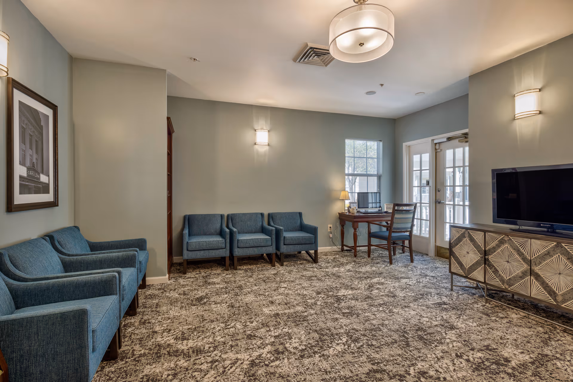 A cozy living room area with blue upholstered chairs arranged along the walls, a patterned carpet, a wooden desk with a chair and lamp near a window, and a TV on a decorative cabinet. The room has soft lighting from wall sconces and a ceiling light fixture.