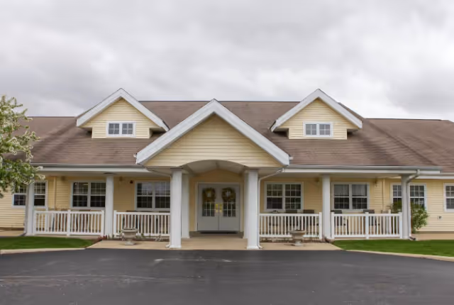 Front exterior view of a single-story senior living facility building with a covered entrance supported by white columns, beige siding, multiple windows, and a paved driveway in front under a cloudy sky.