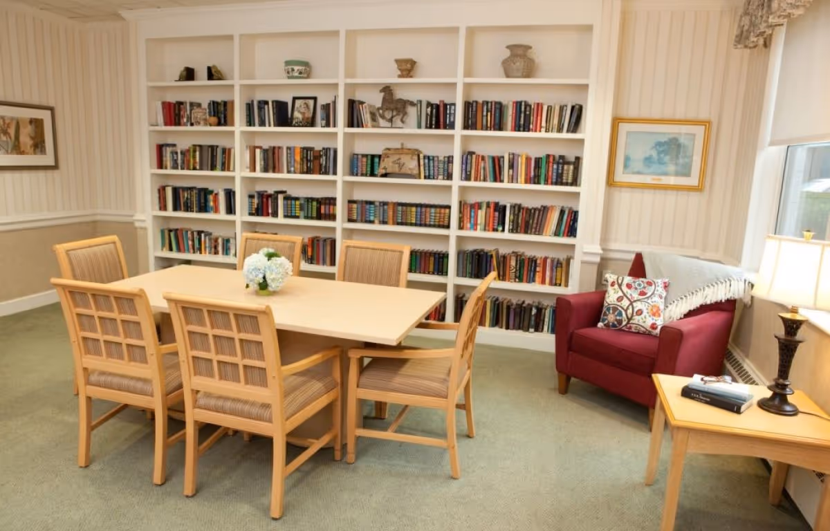 A cozy senior living reading room with a table and six chairs in front of built-in bookshelves and a red armchair by a window.