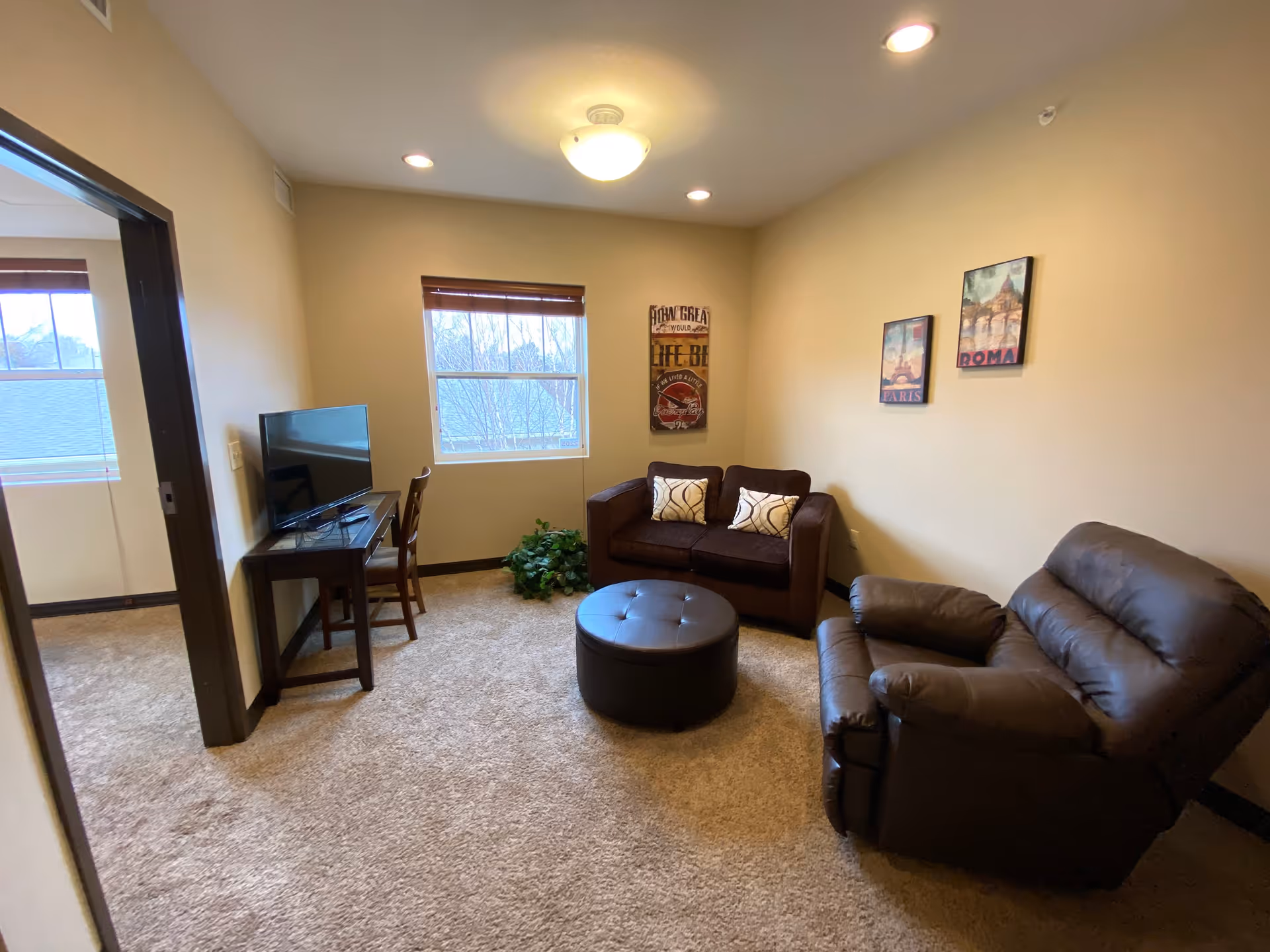 A cozy living room with beige walls and carpeted floor. The room features a brown leather recliner, a dark brown loveseat with two patterned pillows, and a round dark brown ottoman in the center. There is a small wooden table with a flat-screen TV and a chair near a window with wooden blinds. Three framed pictures hang on the walls, and a green plant is placed near the window.