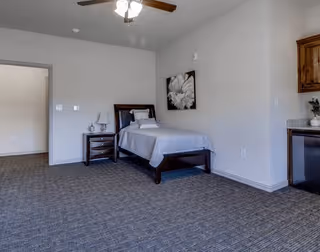 Spacious assisted-living bedroom with a single bed, nightstand and lamp, carpeted floor, ceiling fan, and kitchenette cabinets.