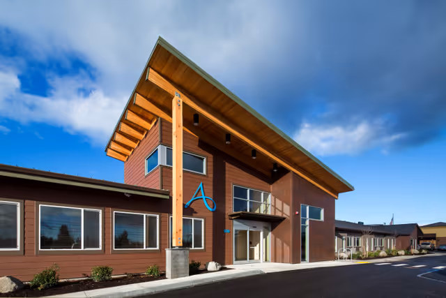 Front exterior of a modern two-story brown building with a large angled wooden canopy over the entrance and windows under a blue sky.