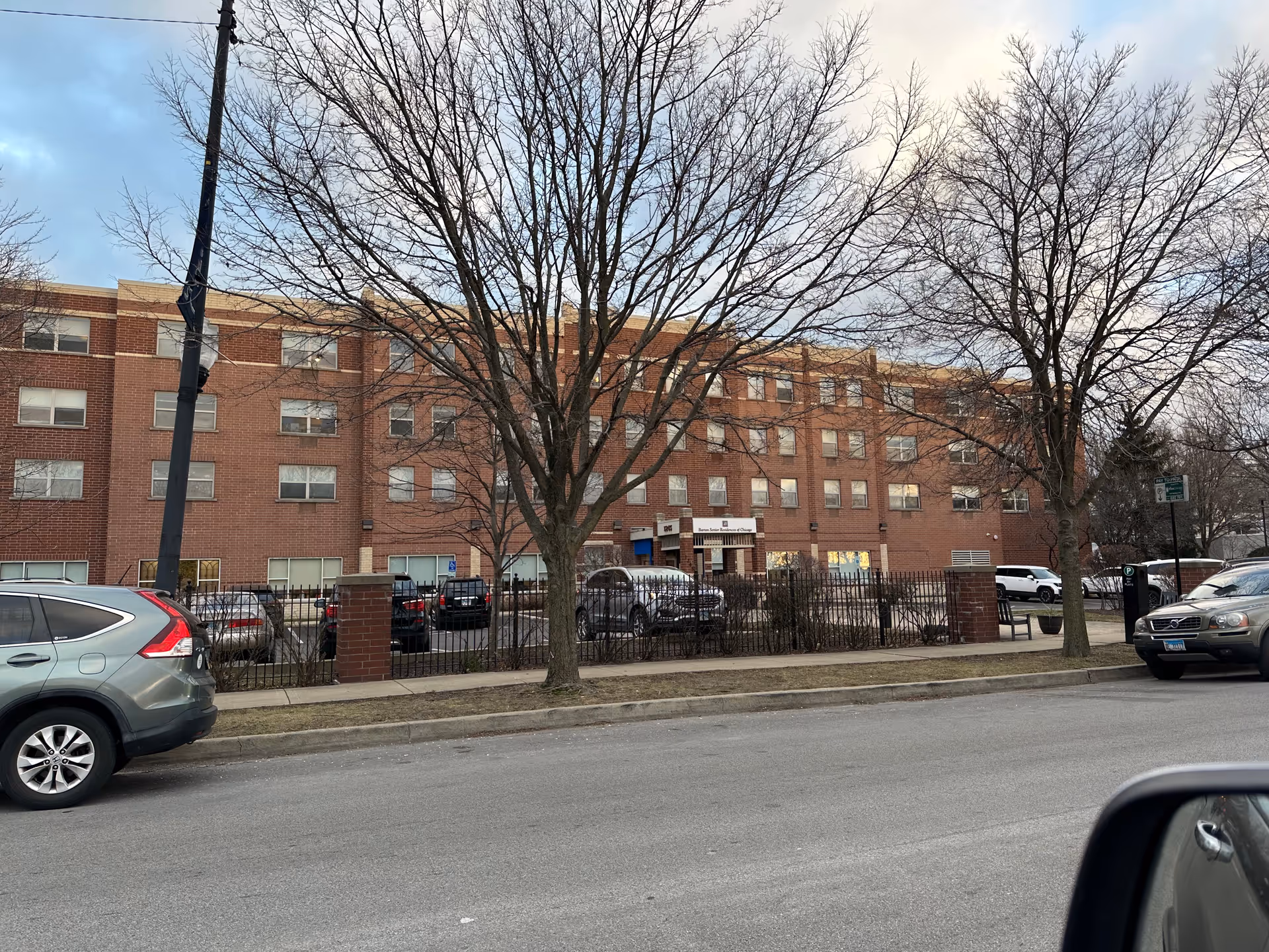 Exterior view of a multi-story brick building with multiple windows, identified as Barton Senior Residences. There are leafless trees and parked cars in front of the building, along with a sidewalk and a street in the foreground.