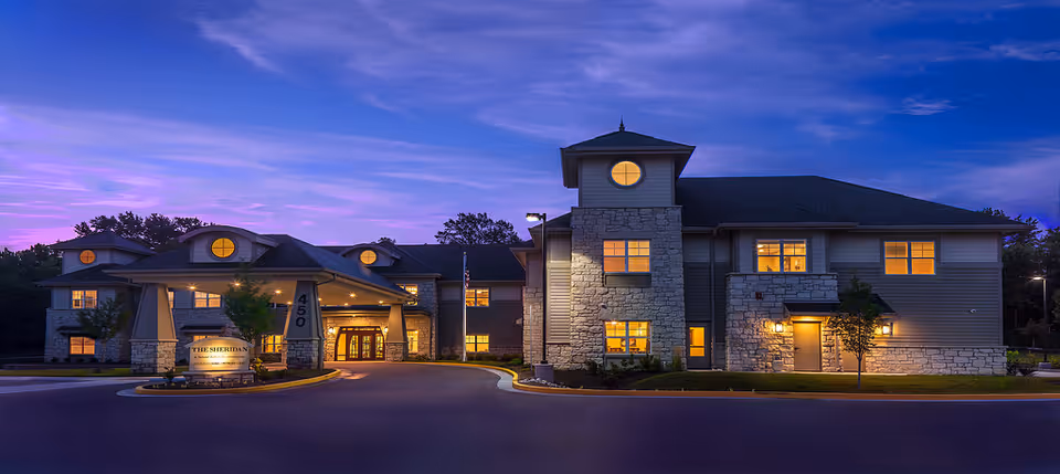 Exterior view of a senior living facility building named The Sheridan at dusk with warm lights glowing from the windows and a clear purple-blue sky in the background.