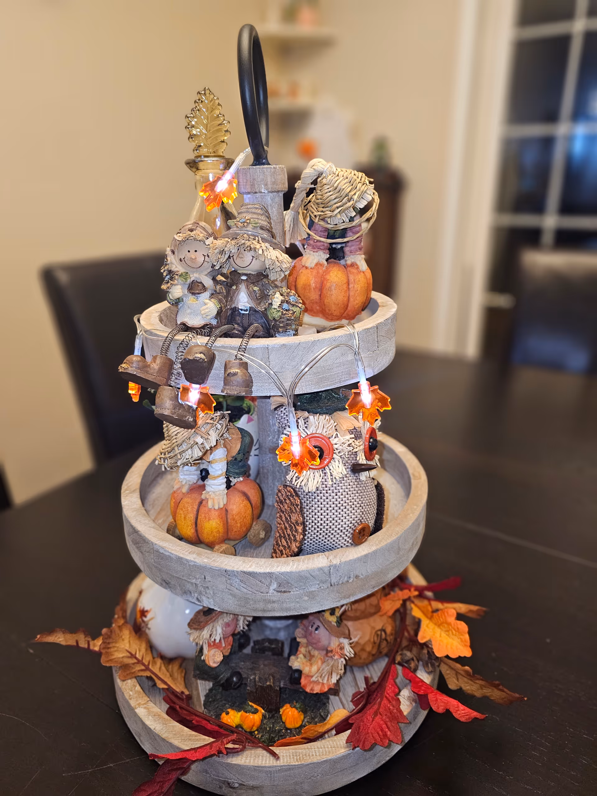 Three-tier wooden centerpiece decorated with autumn pumpkins, owls, scarecrow figurines, and leaf string lights on a dark dining table.