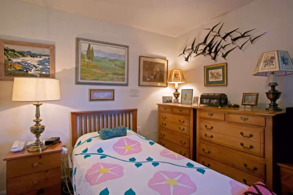 A cozy bedroom with a single wooden bed covered with a floral quilt featuring pink flowers and green leaves. There is a wooden nightstand with a brass lamp and a telephone on the left side of the bed. On the right side, there are two wooden dressers with multiple drawers, decorated with framed pictures, a lamp with a floral lampshade, and a decorative black box. The walls are adorned with various framed paintings and a metal wall art piece of flying birds.