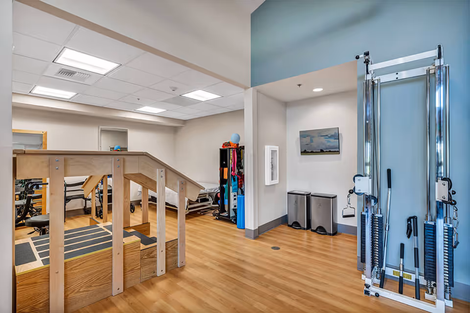 A rehabilitation or physical therapy room with wooden stairs for exercise, a cable machine, exercise equipment, and a treatment table. The room has light-colored walls, wood flooring, and bright overhead lighting.
