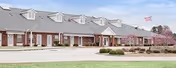 Exterior view of a single-story senior living community building with a brick facade, multiple dormer windows on the roof, and an American flag flying on a flagpole. The building is surrounded by a paved driveway, landscaped bushes, and blooming trees under a clear sky.