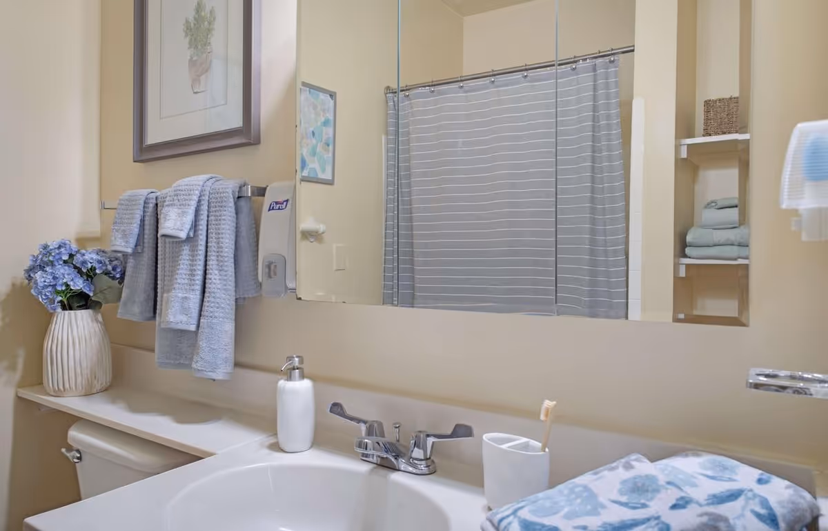Bathroom sink and vanity with a mirror, faucet, soap dispenser, folded towels, shower curtain and a vase of flowers.