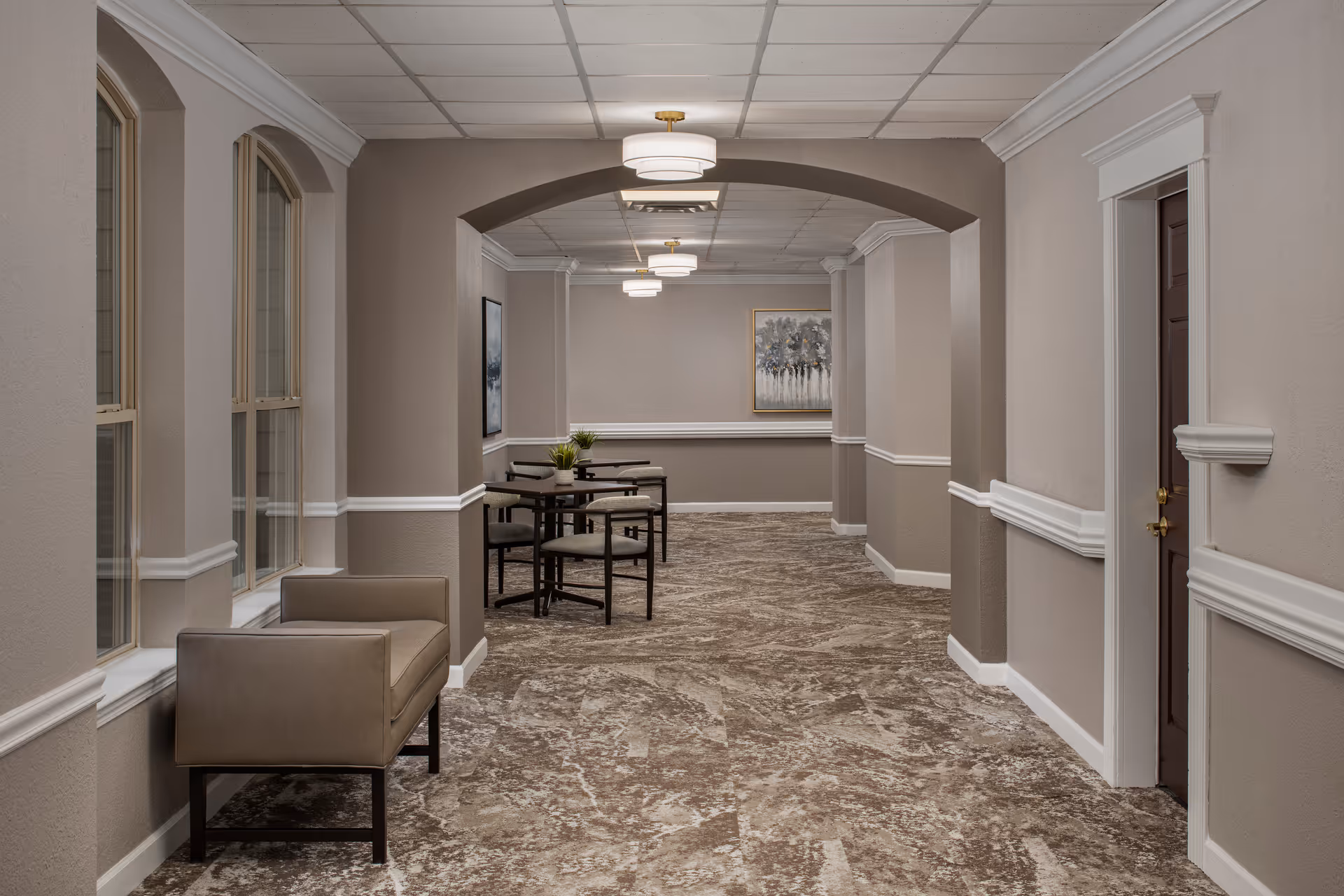 A hallway in an assisted living facility with beige walls, white trim, and a patterned carpet floor. The hallway features arched doorways and windows on the left side, a beige armchair near the windows, and a small seating area with tables and chairs further down the hall. Ceiling lights provide illumination, and framed artwork is visible on the walls.