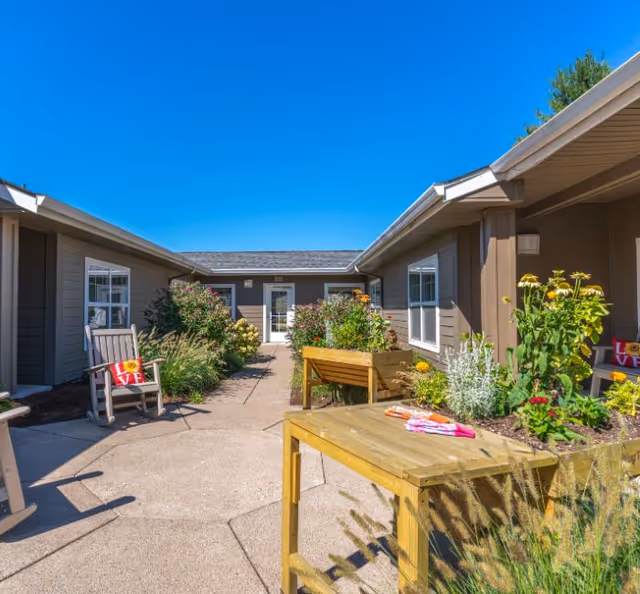 Outdoor courtyard area at Evergreen Senior Living Eugene with a clear blue sky, featuring a wooden rocking chair with a red pillow that says LOVE, raised garden beds with various plants and flowers, and a wooden table with gardening gloves on it.