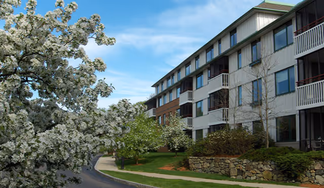 Multi-story residential building with balconies beside a landscaped lawn and blooming white-flowered trees under a blue sky.