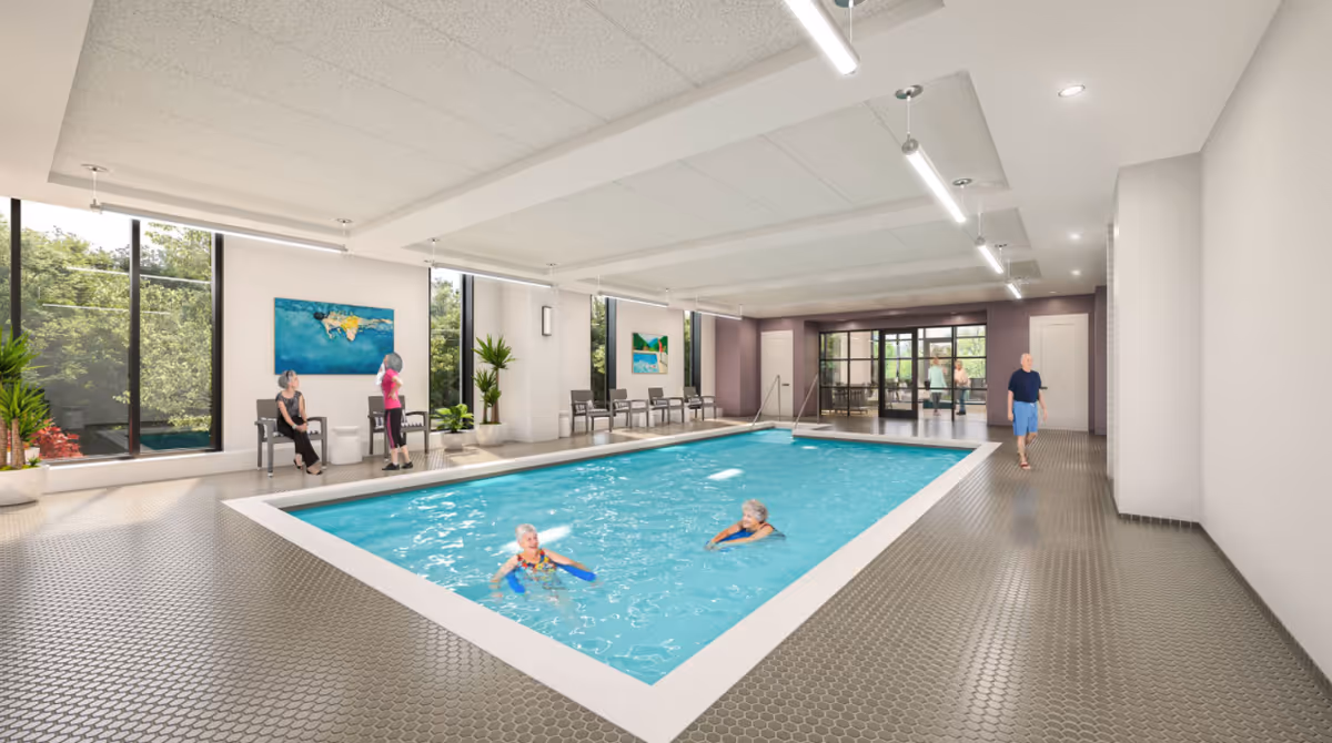 An indoor swimming pool area with large windows, seating along the side, and several seniors swimming and walking nearby.