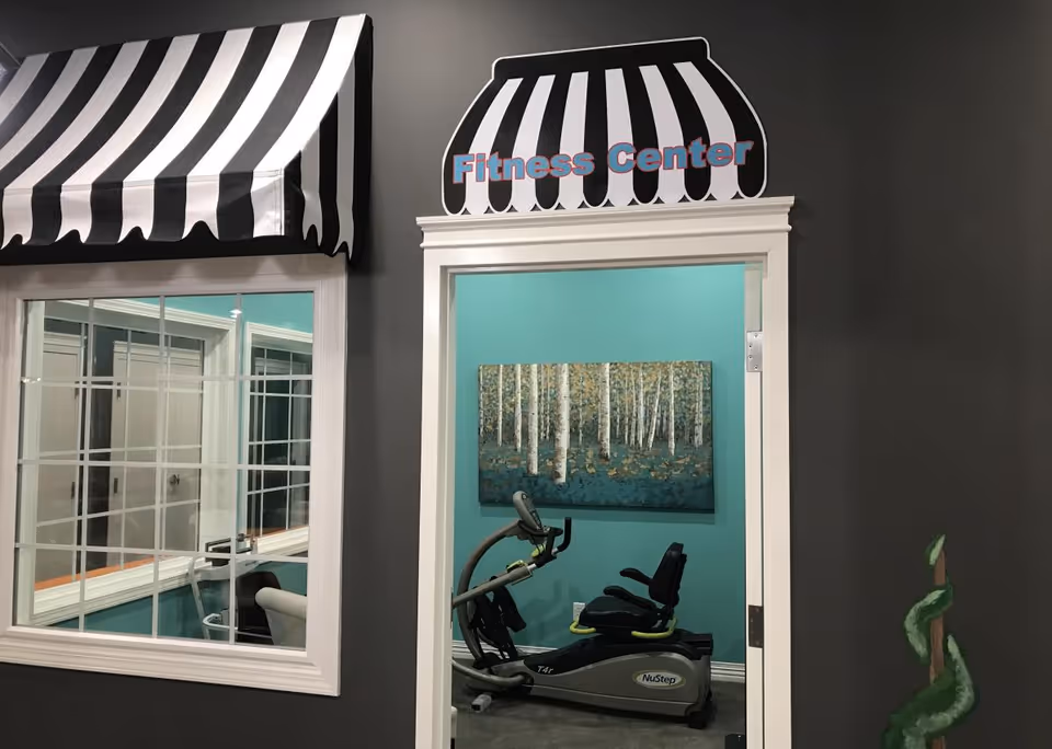 Interior view of a fitness center in a senior living facility with a NuStep exercise machine, teal walls, a painting of birch trees, and a black and white striped awning above the doorway and window.