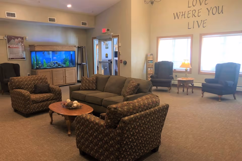 A cozy living room area in a senior living facility with a green sofa, two patterned armchairs, a wooden coffee table with decorative items, two additional armchairs near windows, a side table with a lamp, a large aquarium on a wooden cabinet, and a wall with the phrase 'Love Where You Live'.