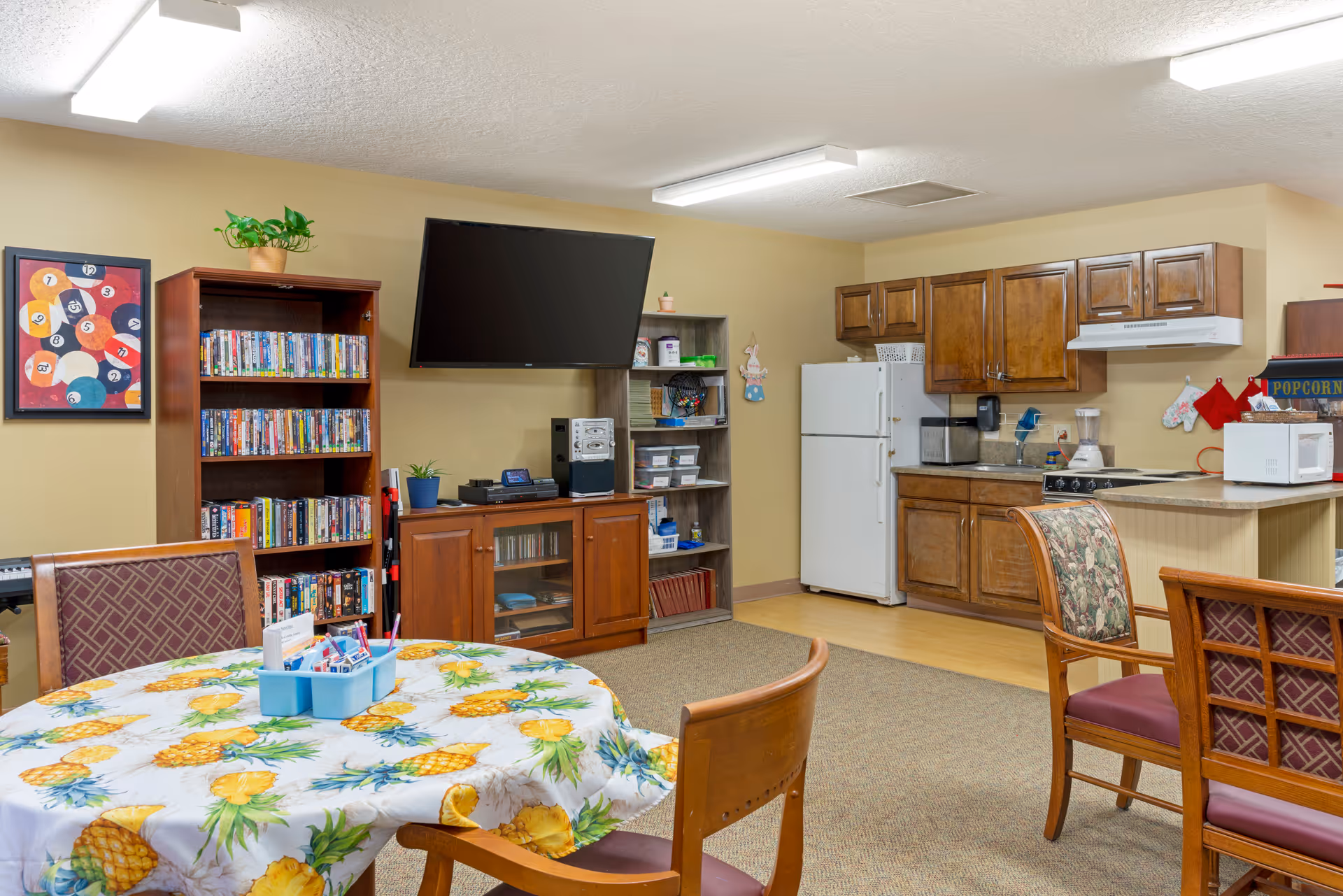 A cozy common area with a round table covered with a pineapple-patterned tablecloth surrounded by chairs. In the background, there is a wooden bookshelf filled with DVDs, a flat-screen TV mounted on the wall, a wooden cabinet with a stereo system, and a small shelving unit with various items. To the right, there is a kitchenette with wooden cabinets, a white refrigerator, a microwave, a blender, and a popcorn machine. The room has beige walls and carpeted flooring.
