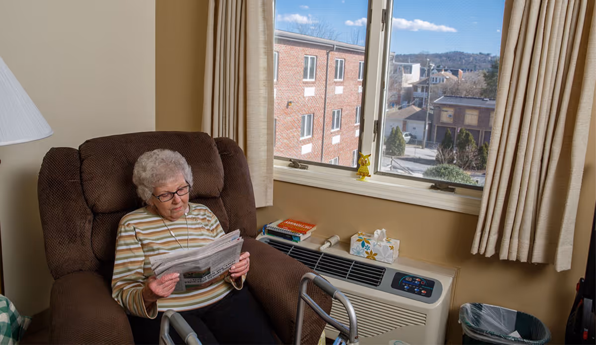 An elderly woman with gray hair and glasses is sitting in a brown recliner chair reading a newspaper. She is wearing a striped long-sleeve shirt. Next to her is a window with beige curtains, a windowsill with books, a tissue box, and a small owl figurine. Outside the window, a brick building and some trees are visible. A walker is positioned in front of her, and a trash can is nearby.