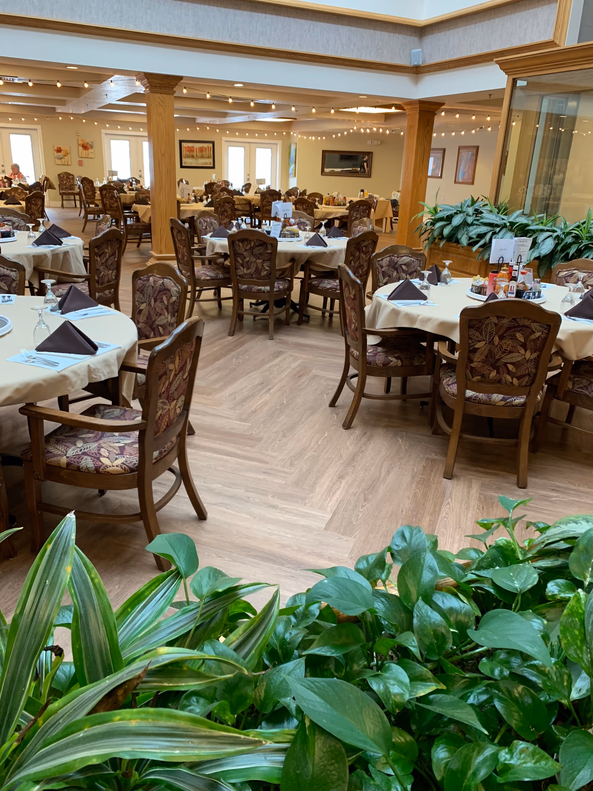 A spacious dining room in an assisted living community with multiple round tables covered with white tablecloths and set with folded brown napkins, glasses, and utensils. The chairs have floral upholstery. There are green plants in the foreground and wooden columns supporting the ceiling. The room is well-lit with natural light from windows and ceiling lights.