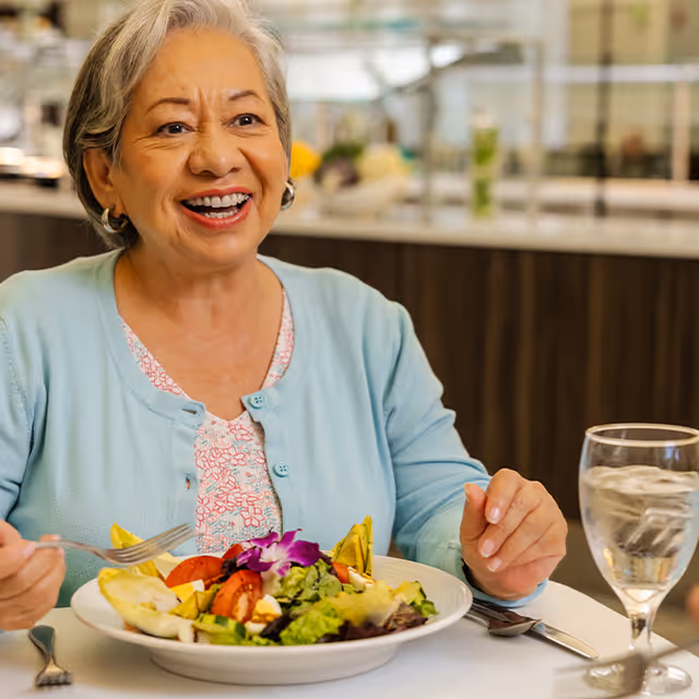 An elderly woman with gray hair wearing a light blue cardigan and floral blouse is smiling while sitting at a table with a plate of salad and a glass of water in front of her in a dining area.
