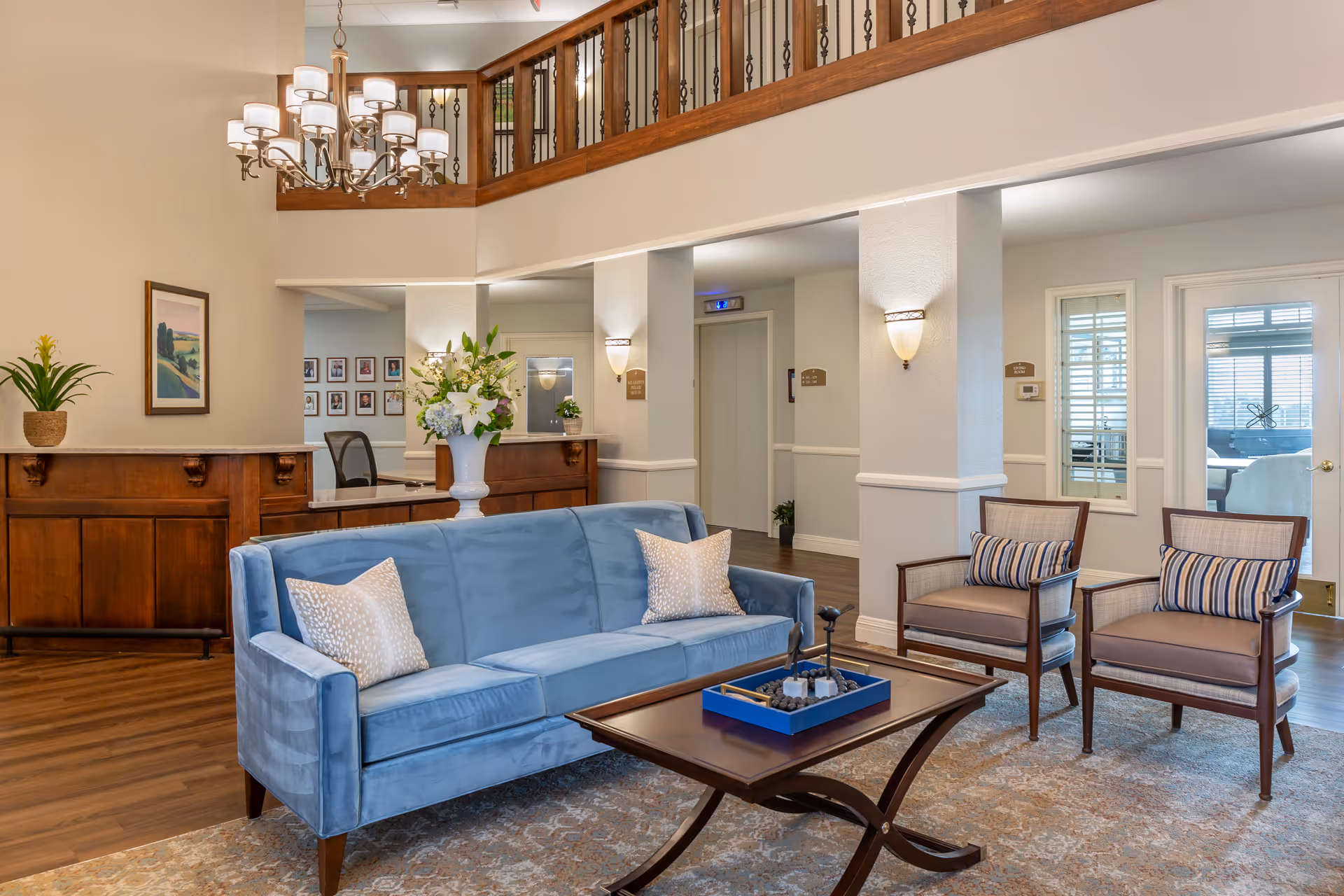 Bright senior living lobby with a blue sofa, two armchairs, a coffee table and a wooden reception desk under a chandelier.