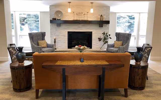 A cozy living room with a brown sofa facing a white stone fireplace. Two gray armchairs with patterned cushions are placed on either side of the fireplace. There are large windows on both sides of the fireplace letting in natural light. Two wooden side tables with decorative vases flank the sofa, and a wooden console table is positioned behind the sofa.